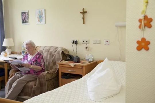 Ältere Person sitzt auf einem Sessel in einem hellen Zimmer mit einem Bett, Nachttisch, Kreuz an der Wand und Blumenbildern.