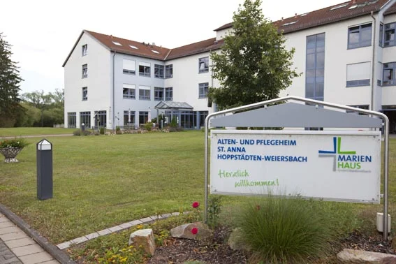 Außenansicht eines mehrstöckigen Alten- und Pflegeheims mit einem Schild, auf dem 'ALTEN- UND PFLEGEHEIM ST. ANNA HOPPSTÄDTEN-WEIERSBACH MARIENHAUS Herzlich willkommen!' steht.