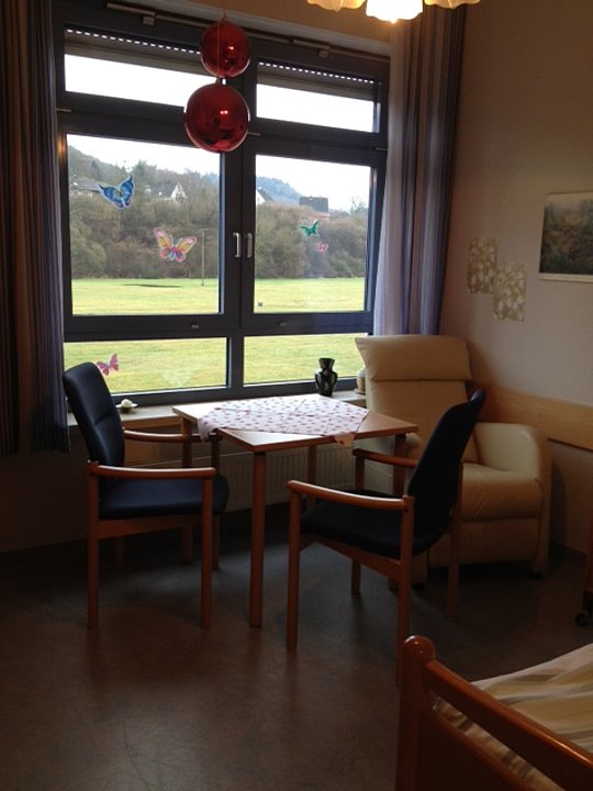 Wohnzimmer mit Fenster, an dem bunte Schmetterlingsaufkleber haften, davor ein Tisch mit zwei Stühlen und ein Sessel.
