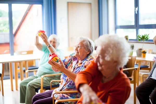 Seniorinnen sitzen in einem hellen Raum auf Stühlen und halten bunte Spielzeuge in die Luft.