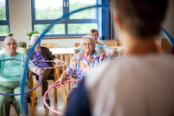Mehrere ältere Menschen sitzen in einem Raum und halten bunte Hula-Hoop-Reifen, während eine Person im Vordergrund ihnen zugewandt ist.